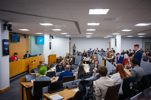 1 WYP member is standing and delivering a speech while the other members listen - in the siambr