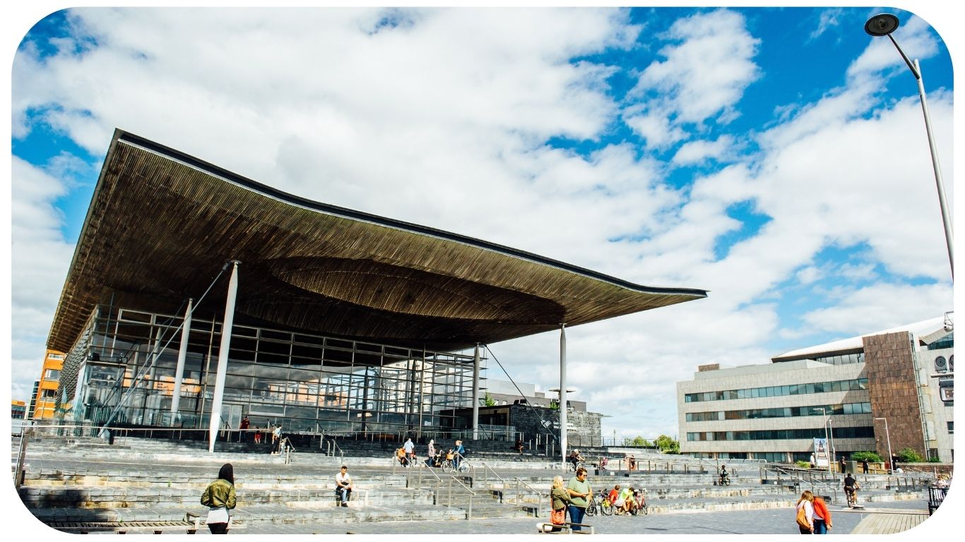 Ffotograff o du blaen adeilad y Senedd ym Mae Caerdydd. Mae'r adeilad wedi'i wneud o frics llwyd, ac mae rhai trawstiau gwyn i'w gweld. Mae arwydd sy'n dweud 'Senedd Cymru'. Yn y blaendir, mae cerddwyr a beicwyr yn mynd heibio. Mae rhai pobl yn eistedd ar y grisiau y tu allan i adeilad y Senedd.