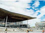 A photograph of the front of the Senedd building in Cardiff Bay. The building is comprised of grey bricks, and some white beams are visible. There is a sign that reads ‘Welsh Parliament’ in the back right hand side. In the foreground, there are pedestrians passing by. Some people are sitting on the steps outside the Senedd building.