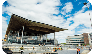 A photograph of the front of the Senedd building in Cardiff Bay. The building is comprised of grey bricks, and some white beams are visible. There is a sign that reads ‘Welsh Parliament’ in the back right hand side. In the foreground, there are pedestrians passing by. Some people are sitting on the steps outside the Senedd building.